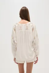 Blusa holgada color blanco roto, con escote en V y mangas largas abullonadas con bordados florales en tono dorado/marrón claro en los hombros y mangas.