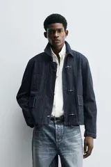 Chaqueta de denim de algodón en color azul oscuro, con cuello solapa y manga larga. Presenta cierre frontal con cremallera oculta bajo una solapa con alamares metálicos, bolsillo de vivo en el pecho y bolsillos de plastrón con solapa en la cadera.