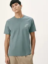 Camiseta blanca de algodón con cuello redondo y mangas cortas. Estampa rectangular con borde negro que contiene la frase "NATURAL ATMOSPHERE" en letras mayúsculas y una imagen de montañas y mar.