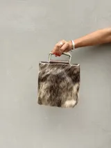 Cartera de mano tipo mini bag confeccionada en cuero con pelo vacuno estampado. Presenta una estructura cuadrada con manija rígida metálica en tono bronce y detalles en cuero liso marrón.