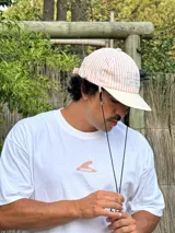 Gorro de visera curva con diseño a rayas verticales en tonos rosado pálido y blanco. La corona tiene un bordado en azul claro con texto. La visera es de color beige claro. Incluye un cordón negro ajustable para colgar del cuello.