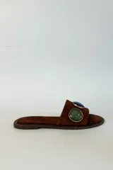 Sandalias planas de gamuza marrón con tira ancha decorada con dos apliques circulares de piedras en tonos verde y azul, enmarcados en metal dorado.
