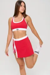 Conjunto deportivo de dos piezas color rojo con detalles en blanco. Incluye un top deportivo con cuello redondo y elástico inferior con logo, y una pollera short con tajo frontal.
