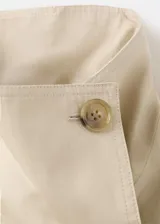 Chaqueta crop estilo gabardina corta color beige, de diseño recto, cuello alto tipo embudo, manga larga, bolsillos laterales y cierre cruzado de doble botonadura con botones oscuros. Con forro interior de poliéster.