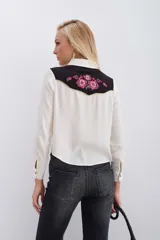 Camisa de estilo western confeccionada en viscosa color crudo. Presenta un canesú delantero y trasero en contraste color negro con bordados florales en tonos fucsia y verde, realzado con vivos en lurex dorado. Cuenta con cierre frontal mediante broches metálicos, mangas largas con puños abotonados y detalles de bolsillos decorativos en el pecho.