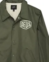 Campera verde militar estilo coach con cuello clásico, cierre con botones a presión, bolsillos laterales y puños elásticos. Estampado con el logo de la marca Deus Ex Machina en el pecho.