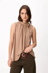 Blusa sin mangas color beige, con cuello alto y detalle de frunces en el frente. Presenta un corte holgado y fluido.
