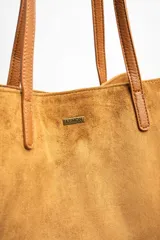 Bolso tipo tote de gran tamaño confeccionado en gamuza sintética color tostado. Presenta un diseño minimalista con asas largas de hombro y una pequeña placa metálica frontal con el logo de la marca.