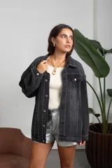 Campera de jean color negro lavado, de corte oversize, con detalle de tachas metálicas plateadas dispuestas en líneas verticales a lo largo de la prenda, cuello clásico, cierre frontal con botones metálicos y bolsillos en el pecho.