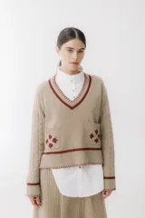 Buzo de punto color visón con escote en V, mangas largas con textura trenzada y dos bolsillos frontales. Presenta detalles de bordados en color bordó en forma de tréboles y terminaciones en contraste en cuello, puños y ruedo.