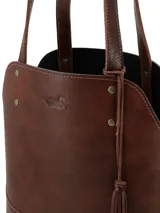 Matera de cuero color marrón con diseño estructurado, costuras laterales decorativas en color claro, base reforzada con remaches metálicos y detalle de borla colgante. Incluye correas largas para llevar al hombro.