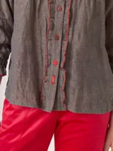 Camisa de lino rústico color gris oscuro con textura visible, con detalles de volados en el frente y pespuntes en hilo rojo. Presenta botones marrones y un pequeño tag rojo con la marca en la vista.