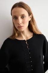 Chaqueta de tejido de punto color negro, con cuello redondo y cierre frontal mediante alamares metálicos. Presenta mangas largas con puños rectos y un corte recto.