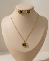 Set compuesto por un collar con cadena fina y dije circular, y un par de aros tipo botón a juego. Ambas piezas presentan una piedra natural ojo de tigre engarzada en una estructura de metal dorado con borde trenzado.