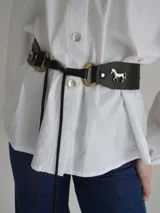 Cinto ancho de cuero marrón con apliques metálicos decorativos en forma de sol, caballo y herraduras, acompañado de tachas.