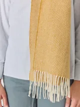 Bufanda de gran tamaño en color mostaza con diseño de tejido en espiga y flecos en los extremos. Incluye una etiqueta metálica decorativa en una de las esquinas.