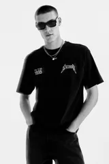 Remera negra de corte holgado con hombros caídos y cuello redondo. Presenta un estampado frontal pequeño con el logo de Metallica y la inscripción 'U.S. Tour 1985'.