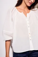 Blusa blanca de algodón peinado, con escote redondo y abertura frontal con botones. Presenta mangas globo de tres cuartos con elástico en el puño y corte holgado.