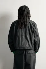 Cazadora regular fit de Zara en colaboración con Aaron Levine, confeccionada en tejido de algodón con acabado efecto encerado en color negro. Presenta cuello subido, manga larga con puño ajustable, bolsillos frontales con solapa y cierre de cremallera oculto con solapa de botones a presión.