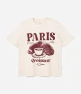 Remera de algodón color crema con cuello redondo y mangas cortas. Presenta un estampado frontal con temática parisina que incluye la palabra 'Paris', un dibujo de una taza de café sobre un croissant y la frase 'Croissant & Crème'.