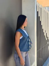 Chaleco de jean azul con cierre frontal mediante lazos ajustables, sin mangas y corte recto.