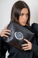 Sobre de mano confeccionado en cuero negro con textura granulada. Presenta un aplique central de piedra ágata natural engarzada en metal dorado. Cuenta con cierre trasero y diseño elegante.