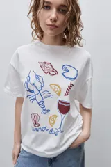 Remera color tostado de corte holgado, con cuello redondo y mangas cortas. Presenta un estampado gráfico frontal de estilo veraniego con ilustraciones de una langosta, conchas, limón, una copa y la inscripción 'Summer Club'.