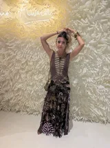 Vestido largo de tul con estampado paisley en tonos marrones, negros y lilas. Presenta un diseño de cuerpo ajustado con cuello redondo y falda con volados en el ruedo. Incluye un detalle de arandela metálica en la cintura y una pieza de joyería dorada superpuesta en el pecho.
