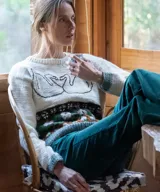 Sweater artesanal de lana merino en color natural, con diseño tejido de jacquard en la parte inferior y bordado a mano de dos cisnes enfrentados formando un corazón en el pecho. Presenta cuello redondo, puños y ruedo en tono verde contrastante.