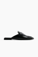Mules estilo mocasín de cuero negro, con punta de almendra y detalle de tira penny con perforaciones decorativas en el empeine. Diseño destalonado con forro de punto.
