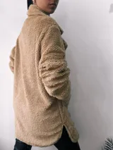Saco de abrigo color beige, de textura suave tipo peluche, con cuello solapa, cierre frontal con botones y bolsillos laterales.