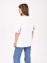 Remera blanca de corte holgado con detalles metalizados en color dorado en las mangas y el ruedo. Presenta un estampado minimalista con texto en el frente.