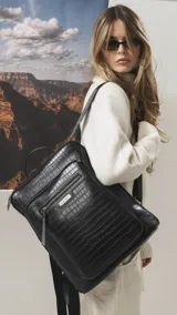 Mochila de diseño rectangular con textura símil cuero efecto cocodrilo en color tostado. Cuenta con un bolsillo frontal con cierre, compartimento principal con doble cierre, correas ajustables y manija superior. Incluye detalles metálicos y placa con logo de la marca.