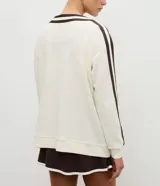 Cardigan deportivo de corte oversized, color blanco con detalles en marrón oscuro. Presenta escote en V, cierre frontal con botones a presión, mangas largas con franjas laterales y puños acanalados. Incluye un parche circular con letras en el pecho.