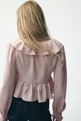 Blusa corta color rosa pálido con tejido suave y leve brillo. Presenta un escote en V pronunciado adornado con un ribete ancho de volados que continúa en el bajo de la prenda. Tiene mangas largas con volumen y puños con volado.