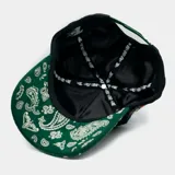 Gorra tipo trucker negra con visera verde estampada con diseño paisley en blanco. Presenta un logo bordado con la letra M en el frente y un bordado detallado de la Virgen de Guadalupe en el lateral.