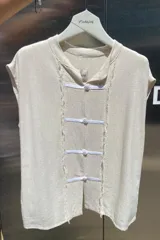 Blusa sin mangas color beige claro, confeccionada en lino tusor. Presenta cuello mao y cierre frontal con tres nudos chinos blancos. Los bordes de la abertura frontal tienen un acabado deshilachado.