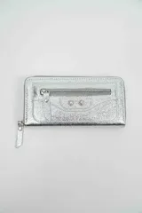 Billetera rectangular de cuero sintético metalizado color plateado, con cierre perimetral y bolsillo frontal con cierre y detalles de tachas metálicas.