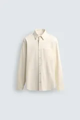 Camisa de algodón color crema o blanco roto, de corte regular fit, con cuello solapa, manga larga con puño abotonado y bolsillo plastrón en el pecho. La tela presenta una textura sutil de rayas verticales en relieve.
