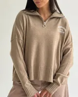 Sweater de tejido bremer color beige, con cuello alto y cierre frontal parcial. Presenta mangas largas con puños acanalados, ruedo acanalado y un bordado decorativo en el pecho.
