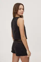 Chaleco corto de denim negro con escote en V y diseño sin mangas. Presenta terminación lavada y detalles laterales con entramado y tachas metálicas que aportan un toque distintivo.