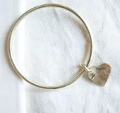 Pulsera tipo brazalete rígido de metal dorado con un dije colgante en forma de corazón que presenta las letras PSM grabadas.