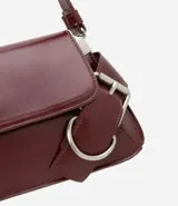 Cartera tipo baguette de tamaño pequeño, confeccionada en poliuretano color negro. Presenta una solapa frontal con detalles de hebillas metálicas decorativas en los laterales y un asa de hombro ajustable.