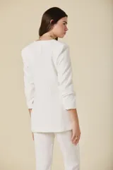 Saco de mujer color crema con diseño estructurado, cierre frontal de un botón dorado y bolsillos laterales con solapa. Presenta mangas tres cuartos con detalle de botones dorados en los puños.