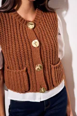 Chaleco tejido color beige con cuello redondo, sin mangas, con cierre frontal de cuatro botones dorados y dos bolsillos frontales.