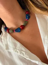 Collar tipo gargantilla compuesto por cuentas de resina en formas irregulares y esféricas, alternando colores rojo, azul y gris jaspeado, separadas por pequeñas cuentas cilíndricas de color fucsia.