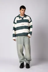 Buzo tipo polo de corte boxy, con diseño de franjas horizontales anchas en color verde oscuro y crema. Presenta cuello camisero, cierre frontal parcial y detalles de costuras desflecadas. Incluye logo bordado en el pecho.