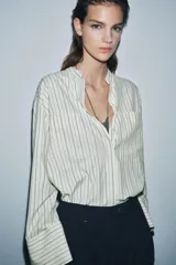 Camisa de corte oversize confeccionada en tejido con viscosa, con estampado de rayas verticales. Presenta cuello mao, cierre frontal con botones ocultos bajo solapa, mangas largas con puños anchos y bolsillos de plastrón en el delantero. El bajo es asimétrico y redondeado.