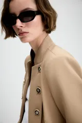 Blazer entallado de sarga color beige con cuello subido y escote en pico. Presenta manga larga con hombreras, detalle de botones metálicos decorativos en el frente y cierre frontal con gancho metálico. Incluye falsos bolsillos de vivo y abertura en la espalda.