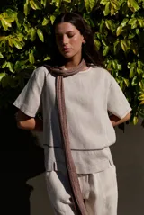 Blusa de lino color beige, de manga corta y cuello redondo, con detalles de costuras decorativas y bordados geométricos a contratono en la parte frontal.
